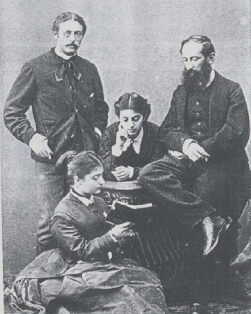 An antique black-and-white studio portrait of a 19th-century intellectual Levantine family; two men and two women in formal Victorian attire, with one woman reading a book. Baltazzi Family