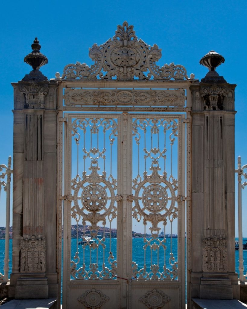 An ornate white sea gate of Dolmabahçe Palace, with its intricate metalwork framing a view of the bright blue Bosphorus.