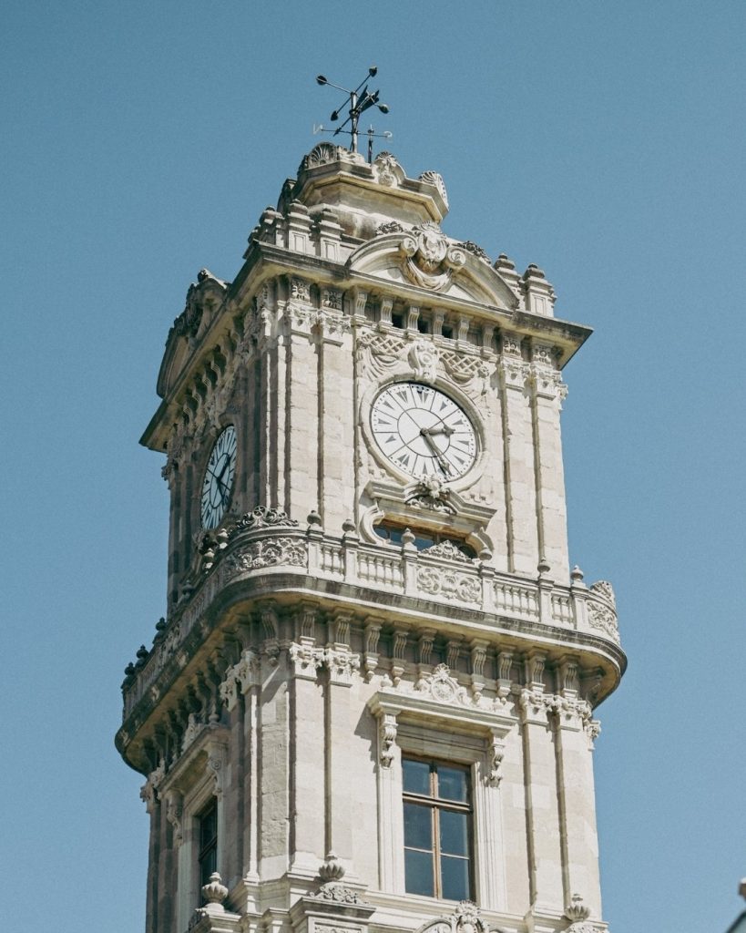 The ornate stone facade of the Dolmabahçe Palace Clock Tower against a clear blue sky, showing the detailed carvings and the clock face.