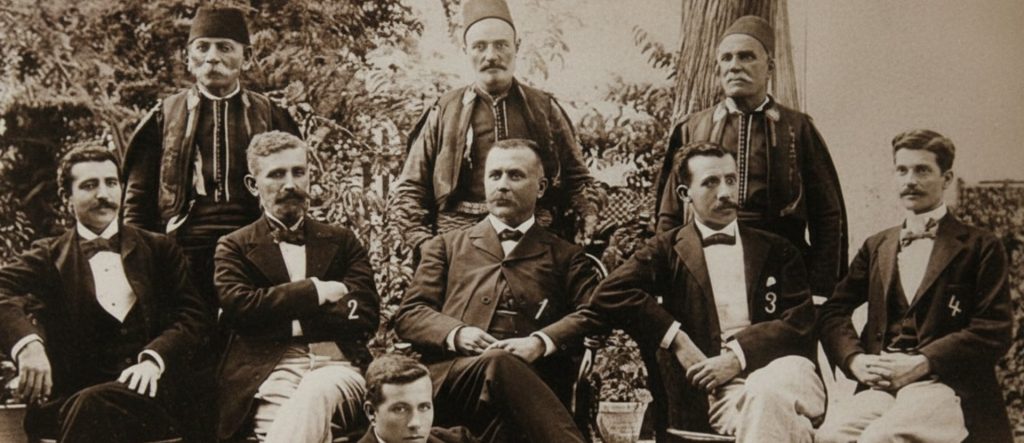 A formal, late 19th-century sepia photograph of a group of men (Galata Bankers), featuring traditionally dressed Ottomans with fezzes standing behind seated men in Western suits, illustrating a blend of cultures.