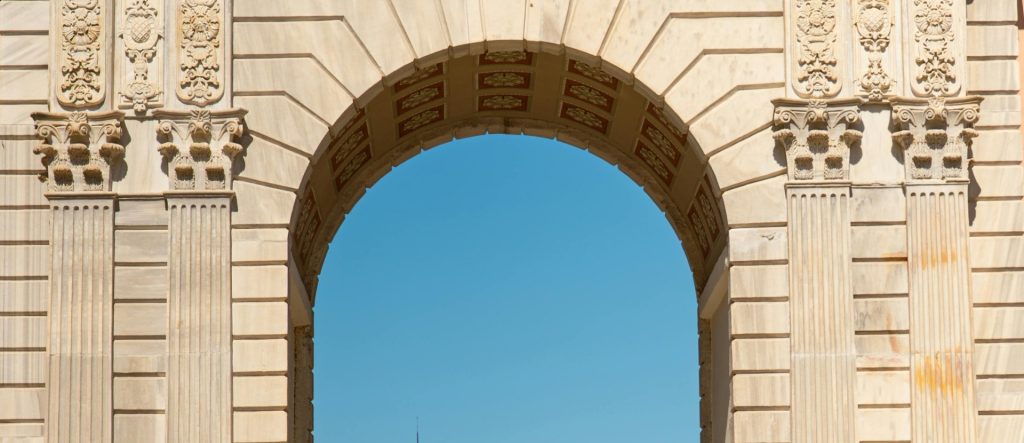 A close-up, panoramic view of a grand stone arch, one of the Gates of Dolmabahce Palace. detailing the intricate Neoclassical carvings and stonework against a clear blue sky.