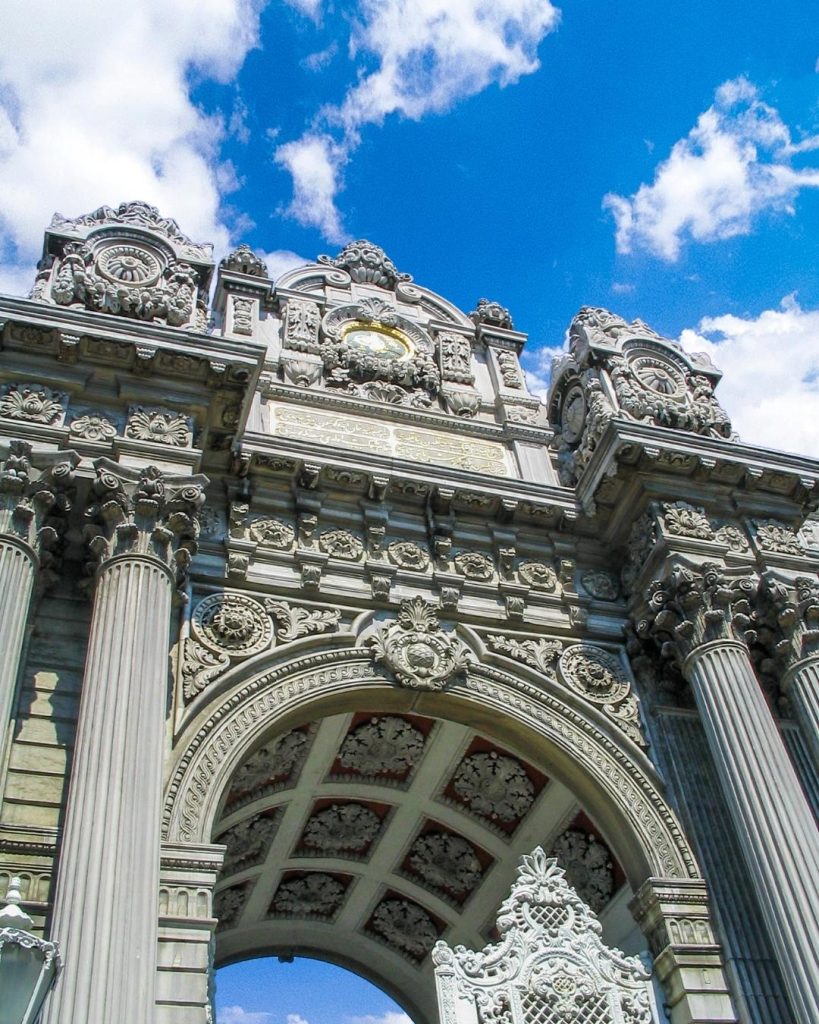 A dramatic low-angle view of the monumental Imperial Gate of Dolmabahçe Palace, showcasing its towering columns and elaborate Baroque carvings under a vivid blue sky with white clouds.