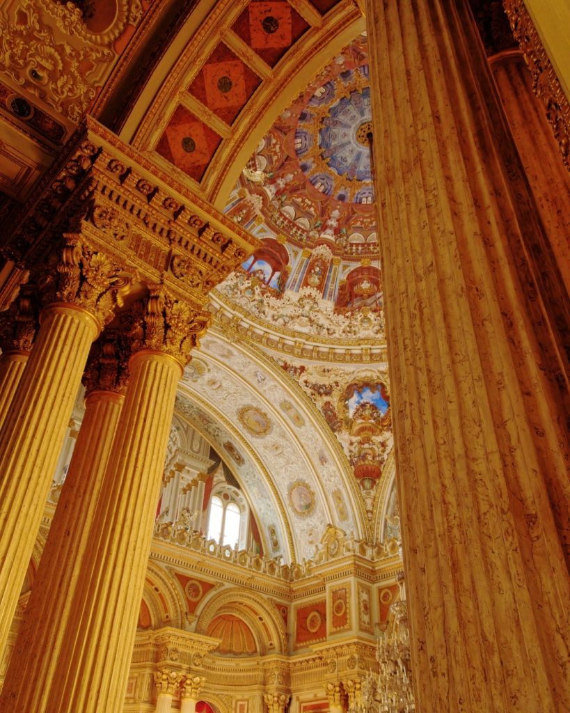 A low-angle view inside the Ceremonial Hall of Dolmabahçe Palace, where majestic gilded Corinthian columns soar towards an intricately painted dome ceiling, capturing the overwhelming opulence of late Ottoman architecture.