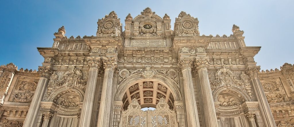 A low-angle, detailed shot of the Imperial Gate (Saltanat Kapısı) of Dolmabahçe Palace, showcasing its highly ornate Baroque and Rococo carvings, towering columns, and the imperial tughra at its crest against a clear blue sky.