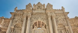 A low-angle, detailed shot of the Imperial Gate (Saltanat Kapısı) of Dolmabahçe Palace, showcasing its highly ornate Baroque and Rococo carvings, towering columns, and the imperial tughra at its crest against a clear blue sky.