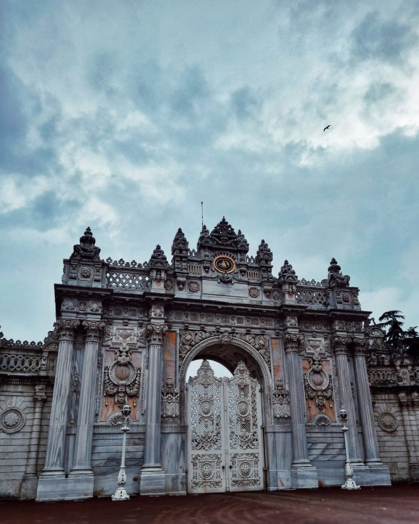 The grand marble facade of the Treasury Gate at Dolmabahçe Palace, standing majestically under a dramatic and moody overcast sky.